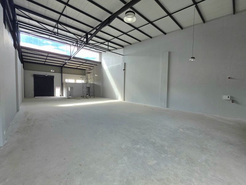 Spacious industrial warehouse unit featuring high ceilings with exposed steel trusses, concrete flooring, and a large roller shutter door for easy access.