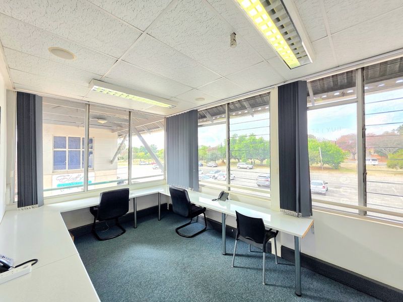 A functional office space featuring built-in wrap-around desks, large louvered windows providing natural light, and a suspended ceiling with integrated lighting.