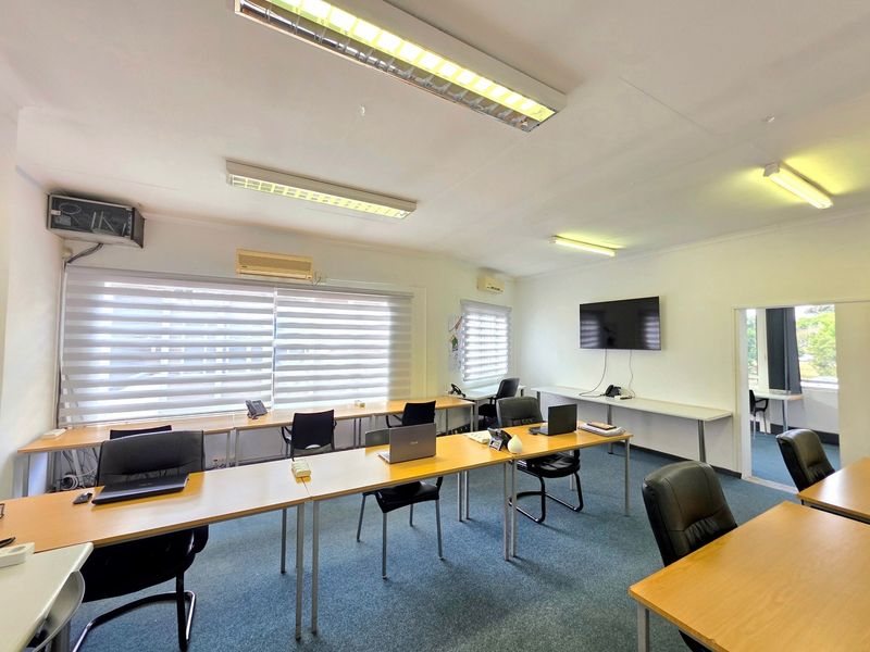 A functional office space featuring carpeted flooring, multiple workstations, wall-mounted air conditioning, and large windows with roller blinds for natural light control.
