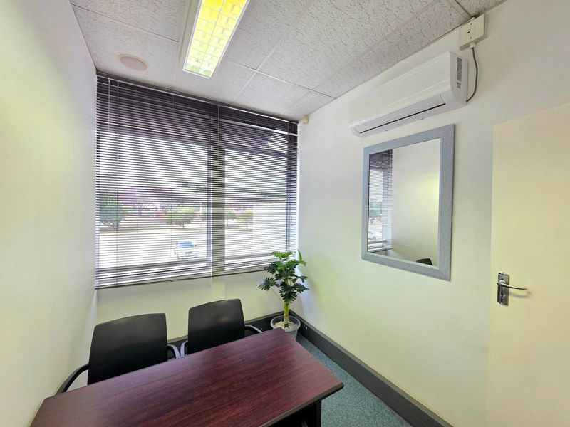 A compact, functional office space featuring a suspended ceiling, split-system air conditioning, carpeted flooring, and large windows with venetian blinds.
