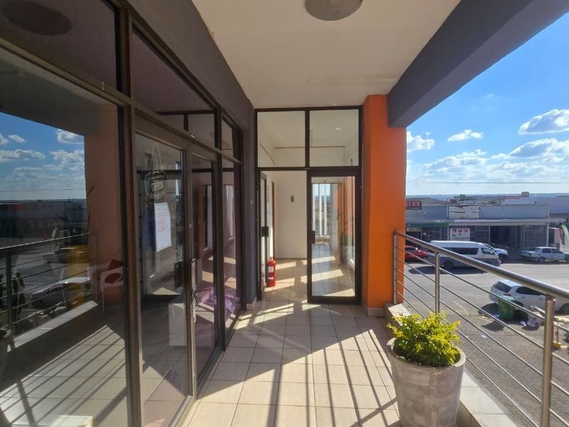 A covered balcony area featuring tiled flooring, stainless steel balustrades, and glass storefront access doors, overlooking a commercial parking area.