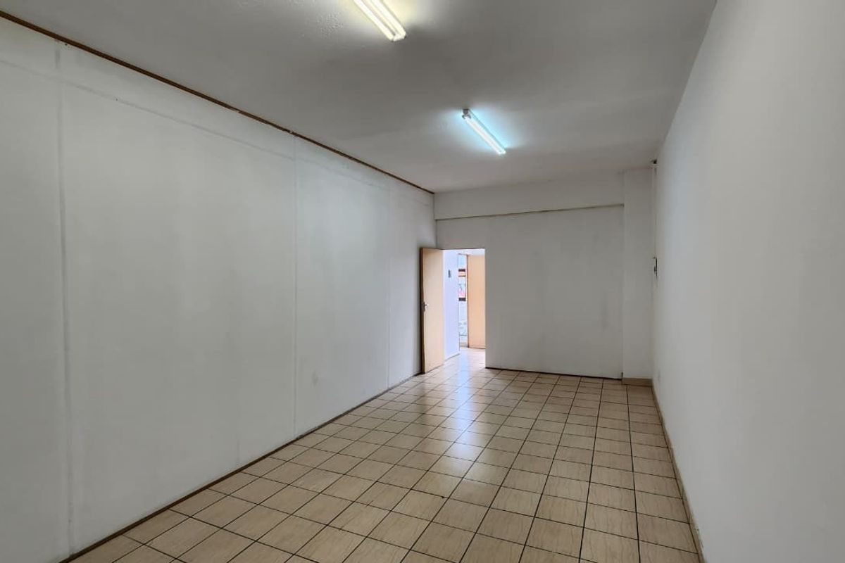 A vacant, rectangular office space featuring neutral white walls, ceramic floor tiling, and overhead fluorescent lighting.