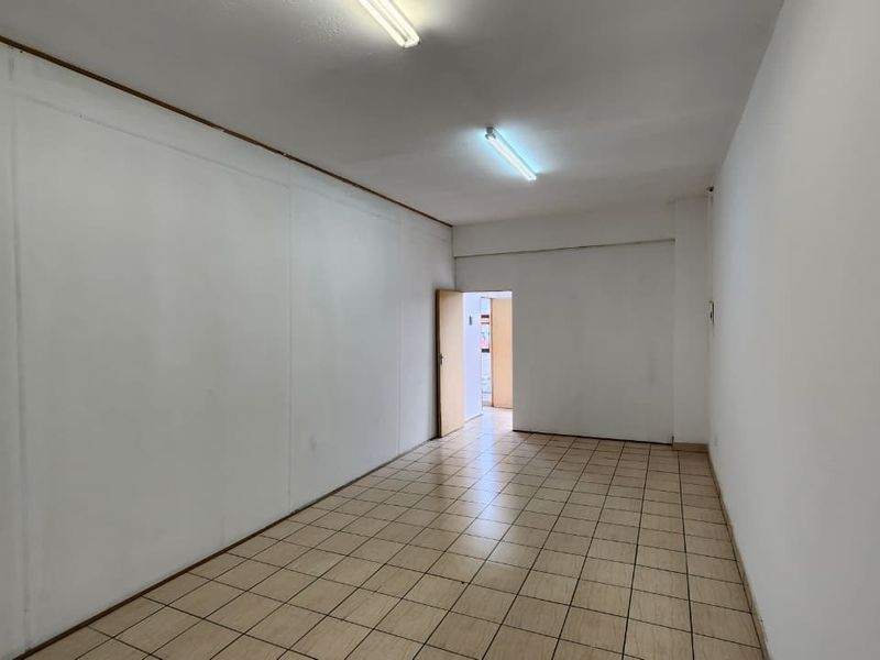 A vacant, rectangular office space featuring neutral white walls, ceramic floor tiling, and overhead fluorescent lighting.