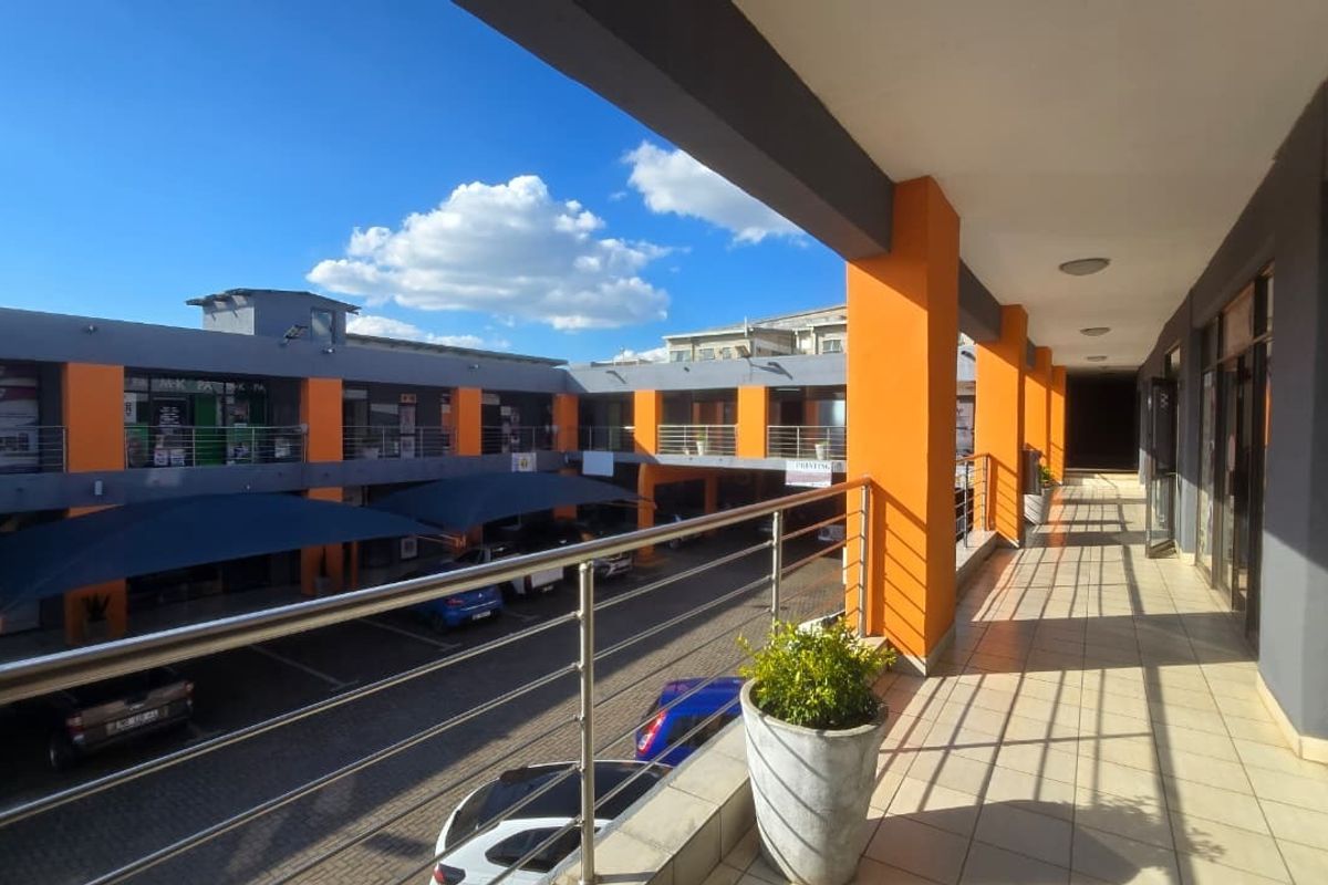 A covered outdoor walkway or balcony overlooking a commercial parking courtyard, featuring tiled flooring and stainless steel railings.