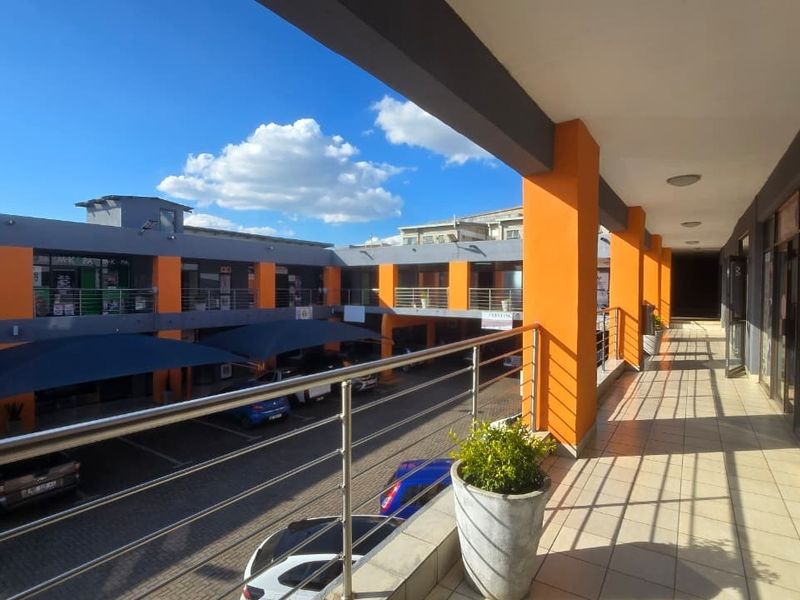 A covered outdoor walkway or balcony overlooking a commercial parking courtyard, featuring tiled flooring and stainless steel railings.