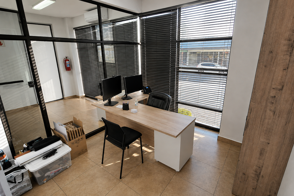 A modern office space featuring glass partition walls, tiled flooring, large windows with venetian blinds, and an integrated air conditioning unit.