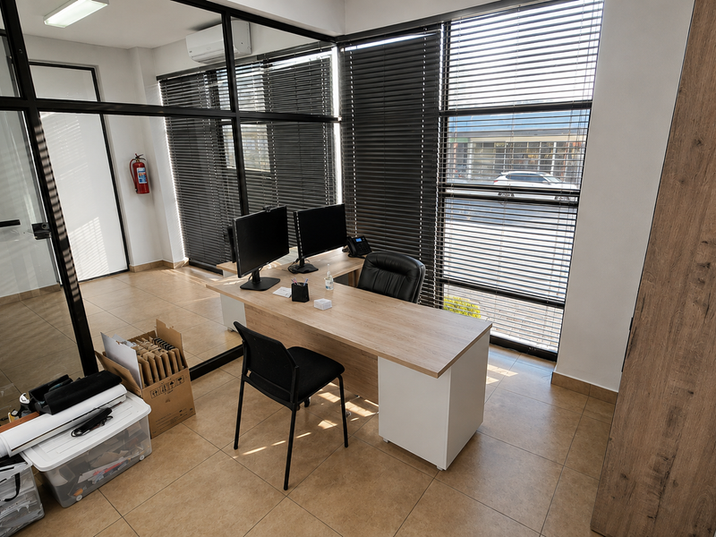 A modern office space featuring glass partition walls, tiled flooring, large windows with venetian blinds, and an integrated air conditioning unit.