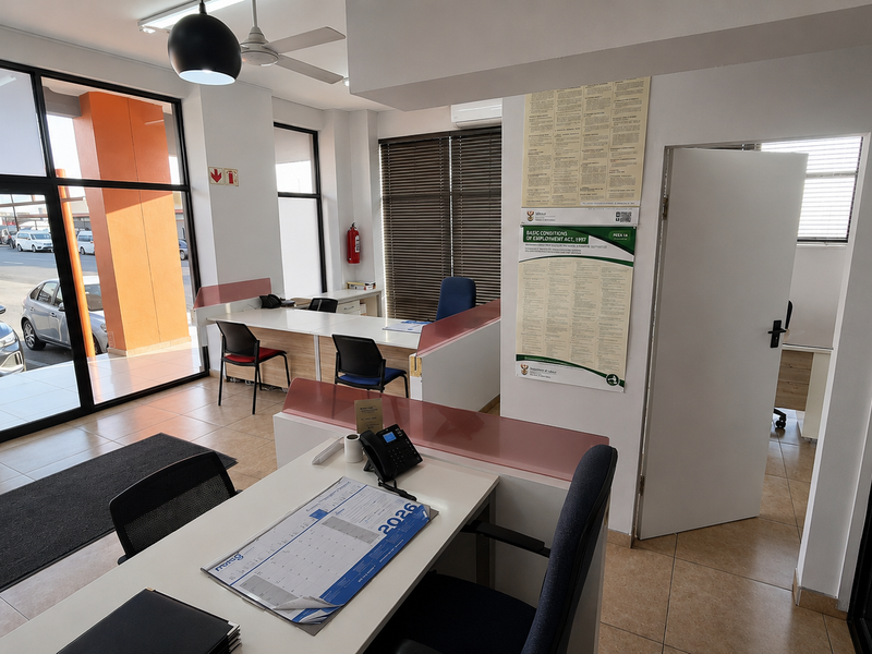 A bright, open-plan office space featuring large glass windows, tiled flooring, built-in desk partitions, and an air conditioning unit.