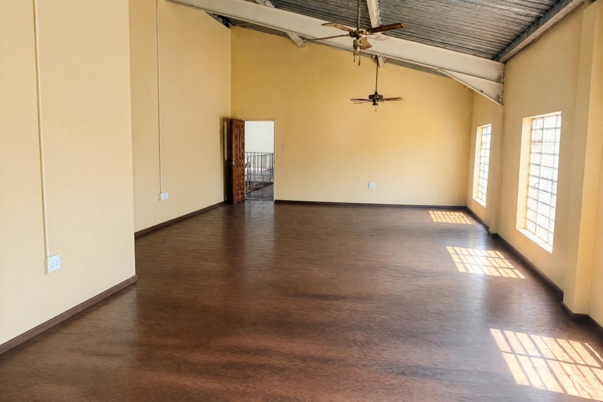 Spacious open-plan office area featuring high vaulted ceilings with exposed metal beams, laminate flooring, and large windows with security bars.