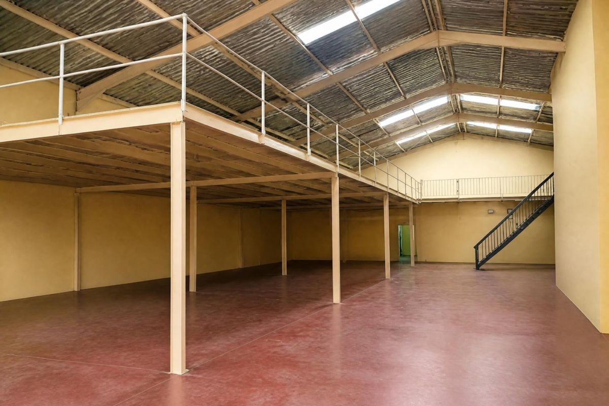 Spacious open-plan office or warehouse area featuring a mezzanine level, steel support columns, metal staircase, and natural light from roof skylights.