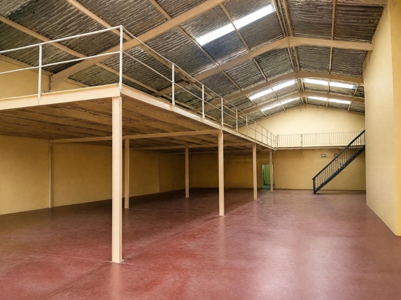 Spacious open-plan office or warehouse area featuring a mezzanine level, steel support columns, metal staircase, and natural light from roof skylights.