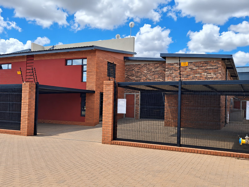 Front exterior view of an industrial property featuring a brick facade, secure gated entrance with electric fencing, and a paved driveway with carport.