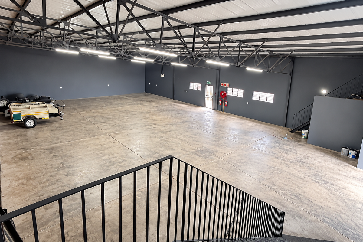 Spacious industrial warehouse unit featuring high ceilings with exposed steel trusses, polished concrete flooring, a brick feature wall, and an internal staircase.