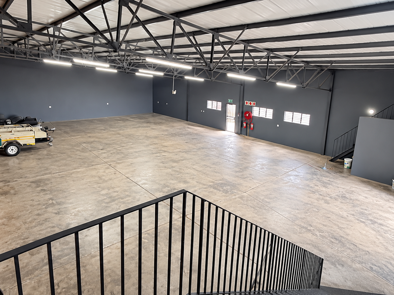 Spacious industrial warehouse unit featuring high ceilings with exposed steel trusses, polished concrete flooring, a brick feature wall, and an internal staircase.