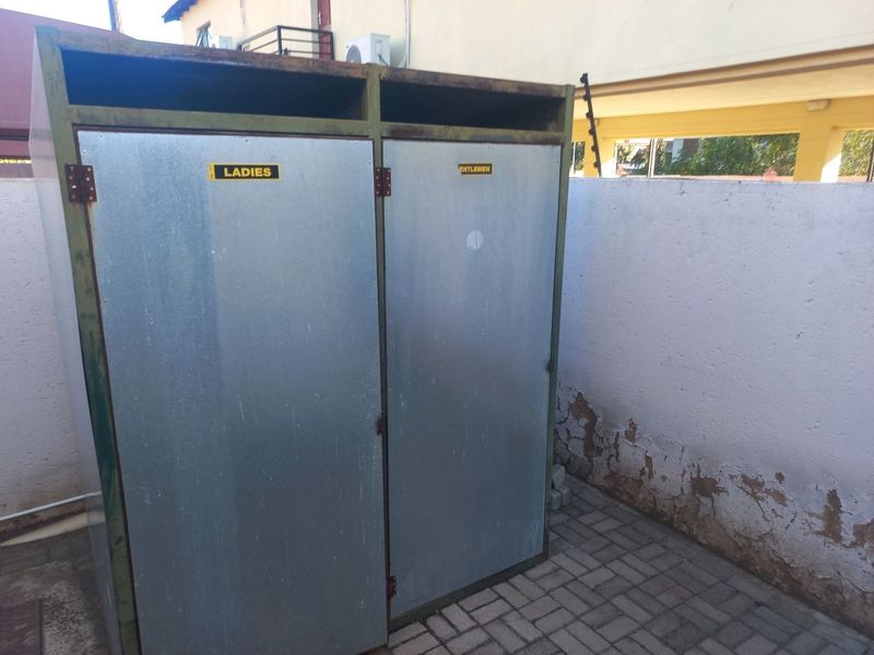 Outdoor metal utility cubicles for ladies and gentlemen, situated on a paved area with a perimeter wall showing signs of wear.