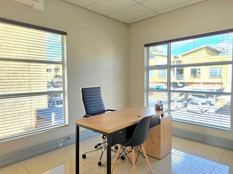A bright, professional office space featuring tiled flooring, large windows with venetian blinds, and a wall-mounted air conditioning unit.