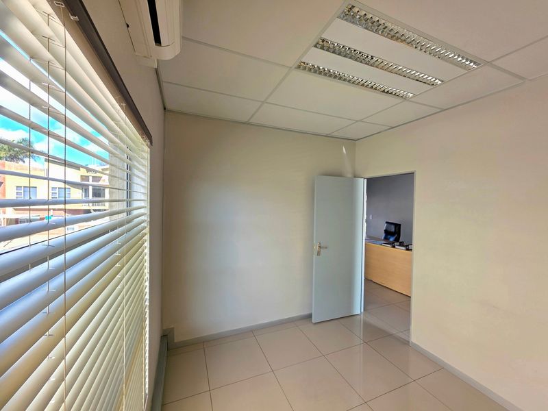 A clean, empty office space featuring tiled flooring, a suspended ceiling with recessed lighting, and a wall-mounted air conditioning unit.