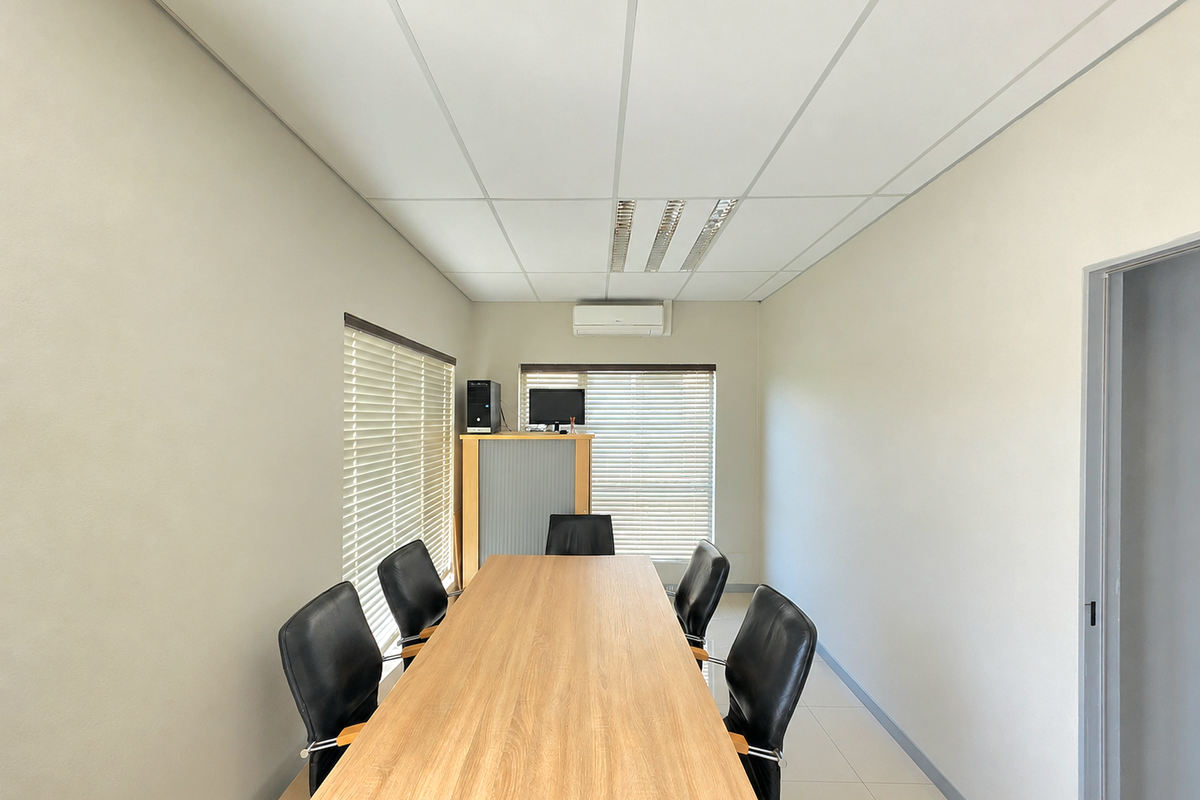 A professional office meeting room featuring a suspended ceiling, recessed lighting, wall-mounted air conditioning, and large windows with venetian blinds.