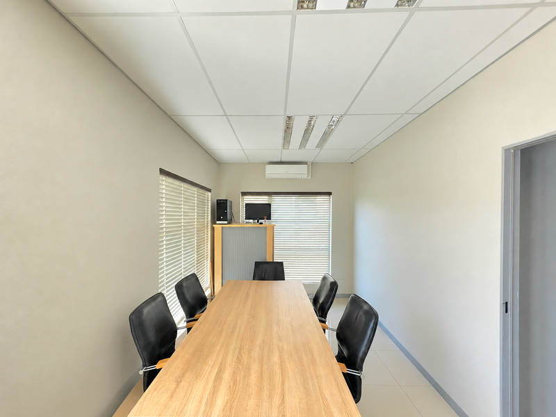 A professional office meeting room featuring a suspended ceiling, recessed lighting, wall-mounted air conditioning, and large windows with venetian blinds.
