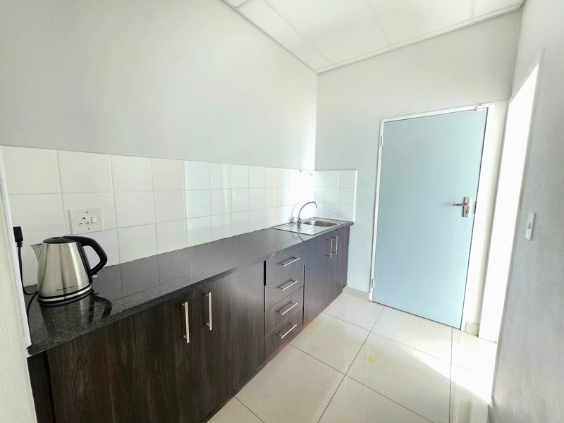 A clean, functional office kitchenette featuring dark wood cabinetry, granite countertops, a stainless steel sink, and white tiled backsplash.