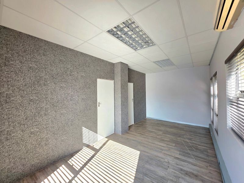 A bright, empty office space featuring laminate flooring, textured wall finishes, a suspended ceiling with recessed lighting, and a wall-mounted air conditioning unit.