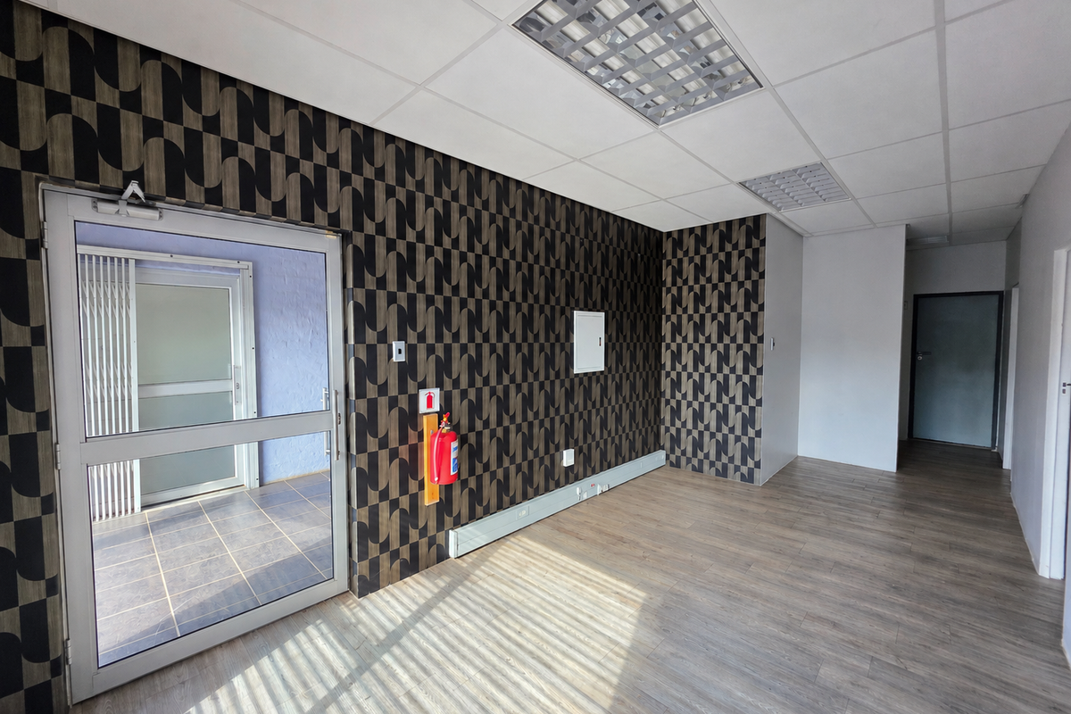 A bright, empty office space featuring modern patterned wallpaper, laminate flooring, suspended ceilings with recessed lighting, and a glass entrance door.