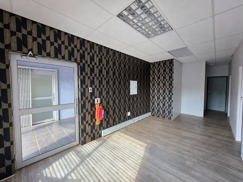 A bright, empty office space featuring modern patterned wallpaper, laminate flooring, suspended ceilings with recessed lighting, and a glass entrance door.