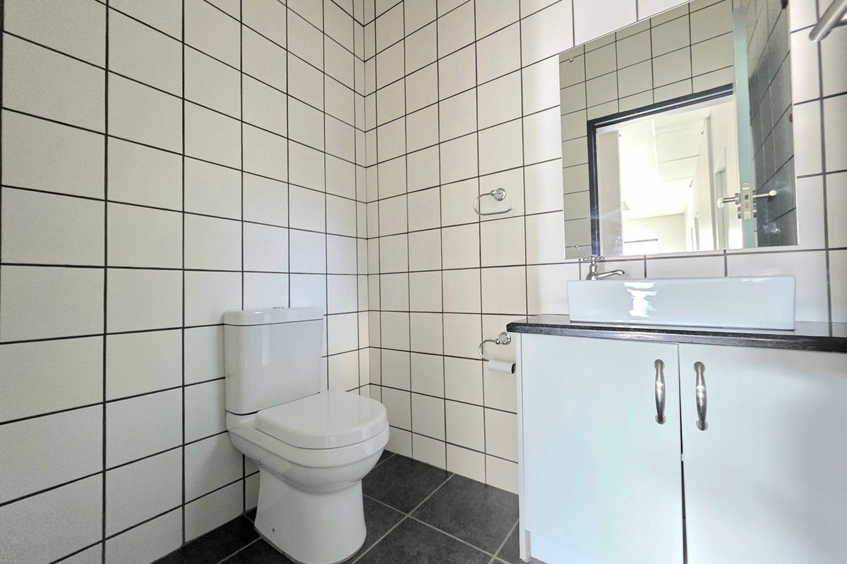 A clean, modern office bathroom featuring white square wall tiles, dark floor tiles, a vanity cabinet with a vessel sink, and a wall-mounted mirror.