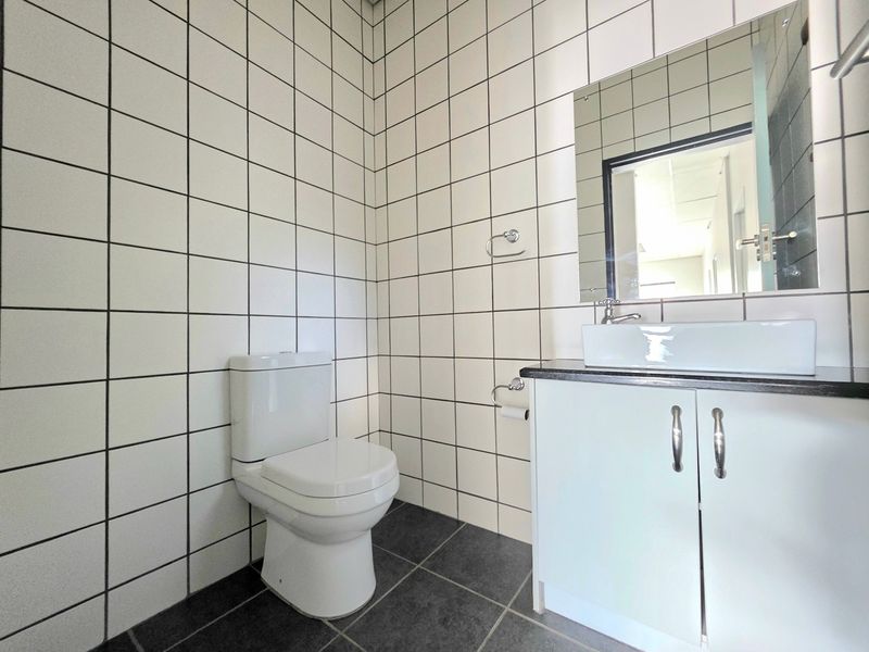 A clean, modern office bathroom featuring white square wall tiles, dark floor tiles, a vanity cabinet with a vessel sink, and a wall-mounted mirror.