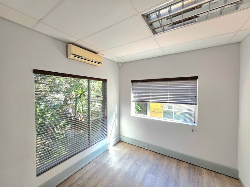 A bright, empty office space featuring laminate flooring, a suspended ceiling with recessed lighting, and a wall-mounted air conditioning unit.