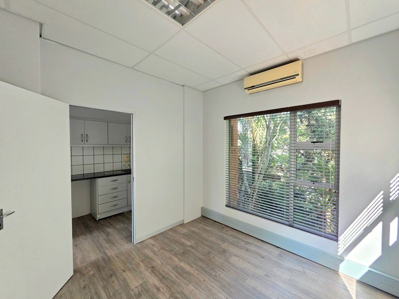 A bright, empty office space featuring laminate flooring, a suspended ceiling, an air conditioning unit, and an adjoining kitchenette area with built-in cabinetry.