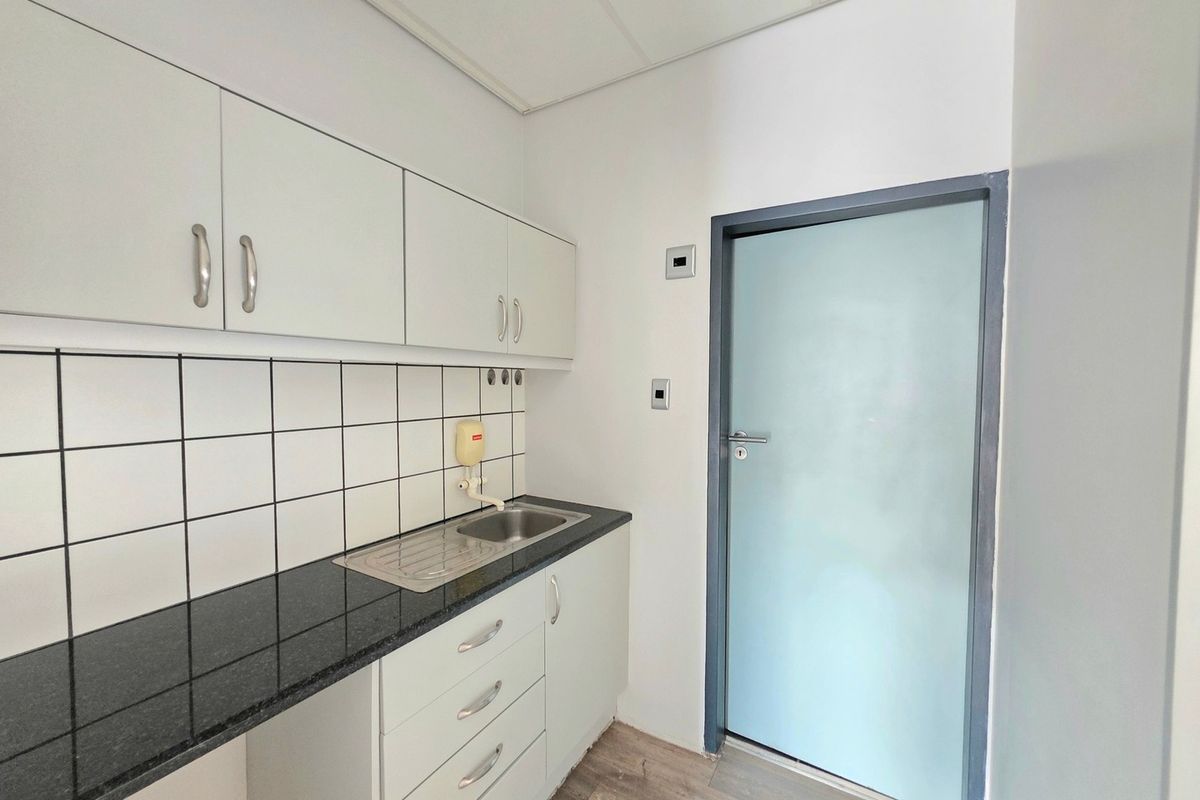 A clean, functional office kitchenette featuring white cabinetry, a dark laminate countertop, stainless steel sink, and a modern frosted glass door.
