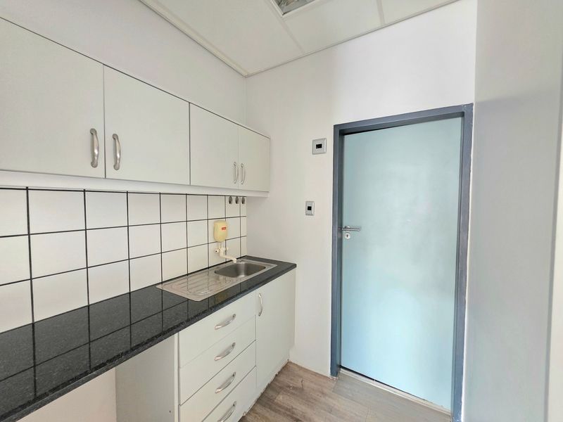 A clean, functional office kitchenette featuring white cabinetry, a dark laminate countertop, stainless steel sink, and a modern frosted glass door.