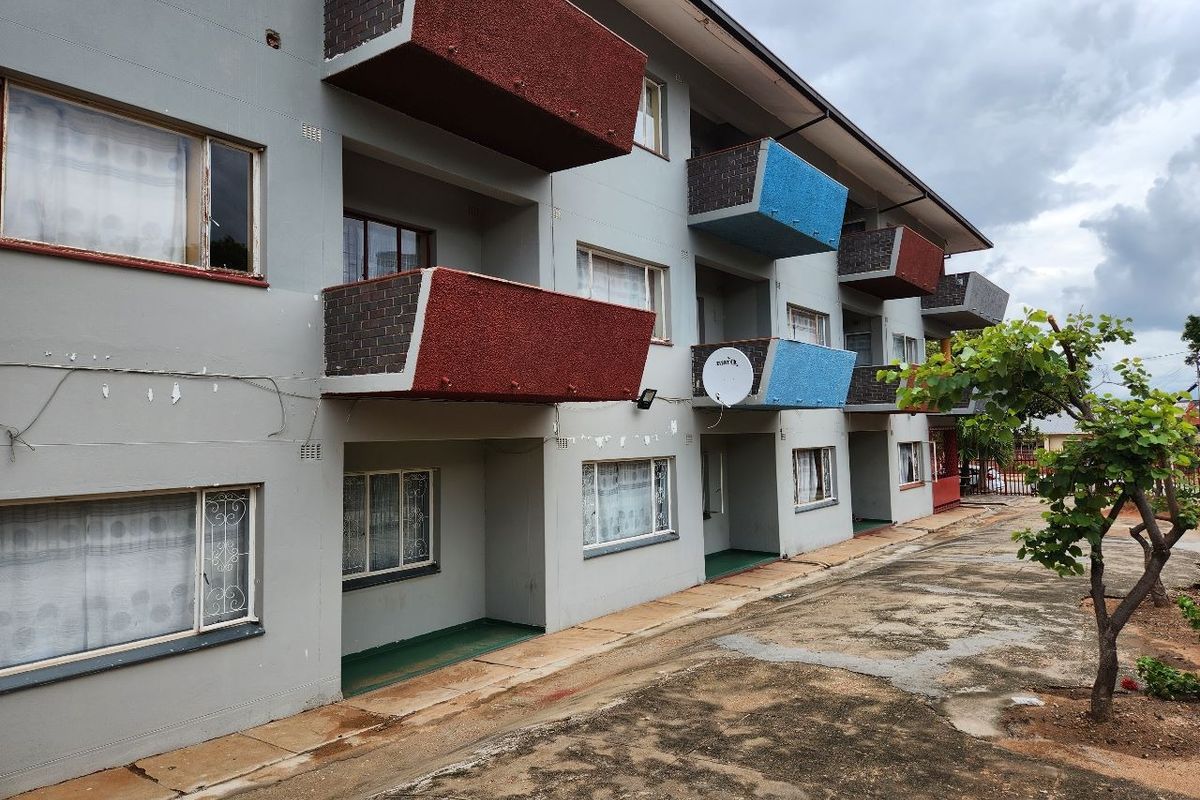 Exterior view of an apartment building featuring multiple balconies, brick facade detailing, and paved ground area.