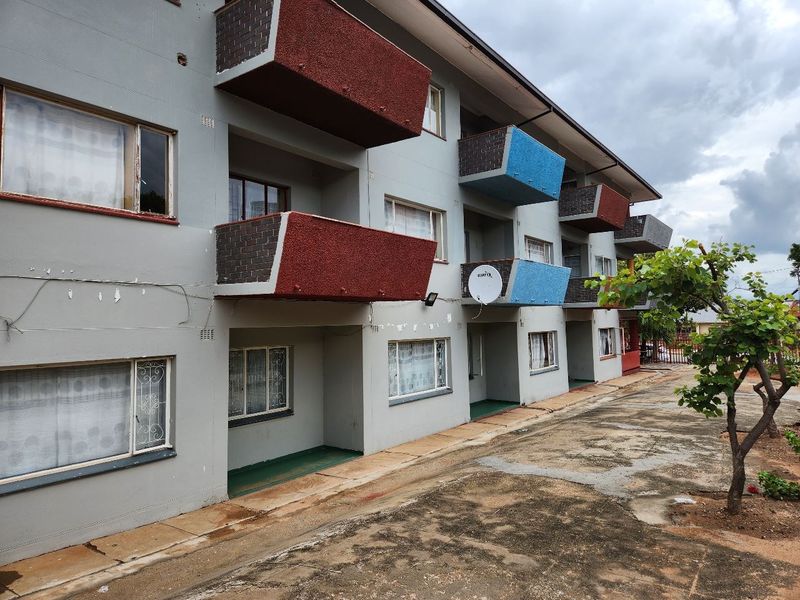 Exterior view of an apartment building featuring multiple balconies, brick facade detailing, and paved ground area.