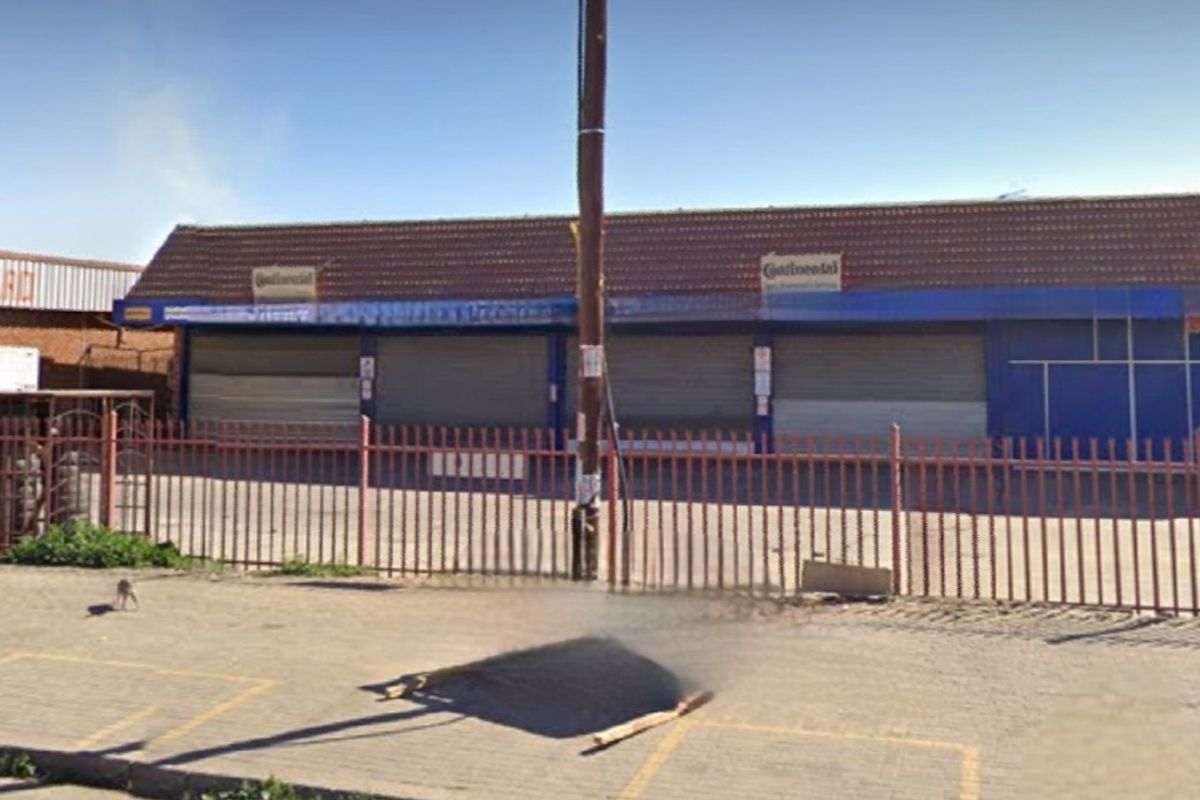 Front elevation of a commercial property featuring a paved forecourt, perimeter steel fencing, and multiple security roller shutters protecting the storefront.