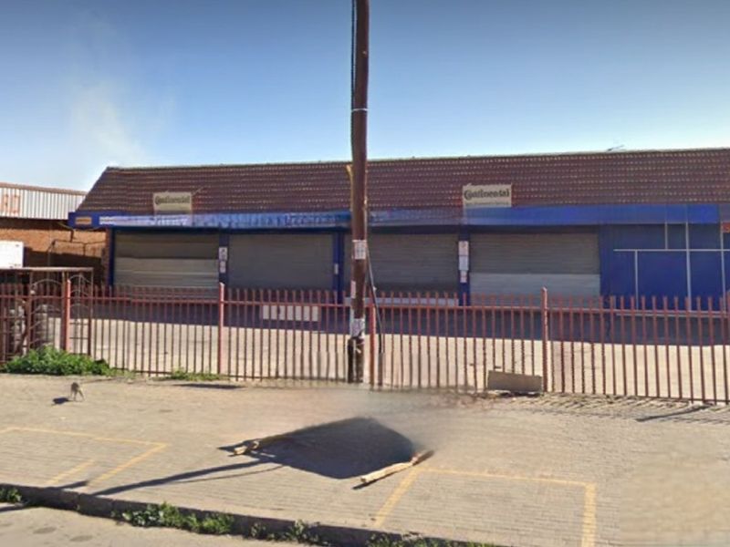 Front elevation of a commercial property featuring a paved forecourt, perimeter steel fencing, and multiple security roller shutters protecting the storefront.