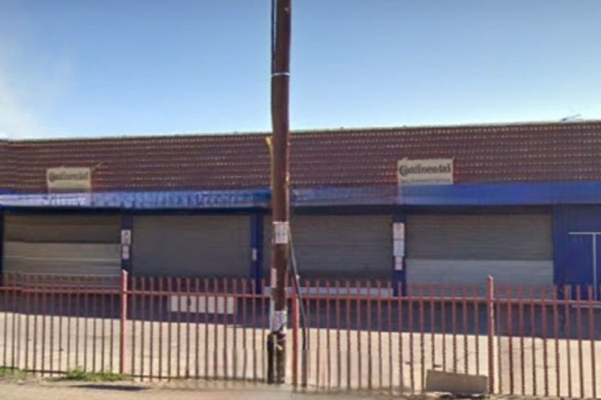 Commercial storefront featuring multiple security roller shutters, a tiled roof, and metal perimeter fencing, showing signs of age and weathering.