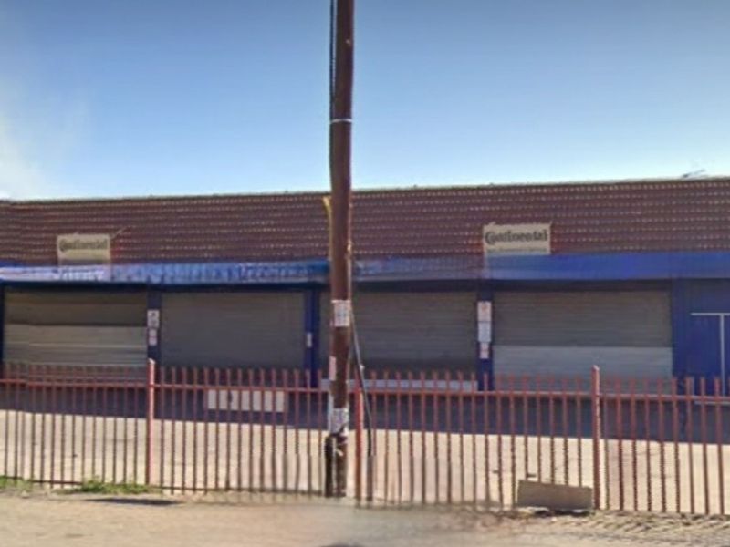 Commercial storefront featuring multiple security roller shutters, a tiled roof, and metal perimeter fencing, showing signs of age and weathering.