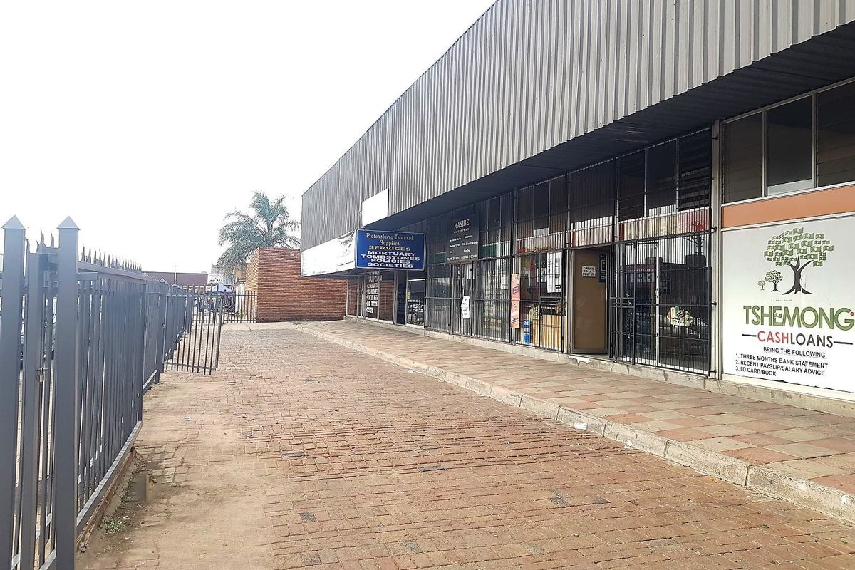 Exterior view of a commercial storefront featuring a paved walkway, metal security fencing, and corrugated roof overhang with signage.