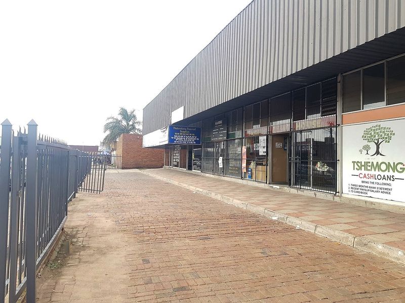 Exterior view of a commercial storefront featuring a paved walkway, metal security fencing, and corrugated roof overhang with signage.