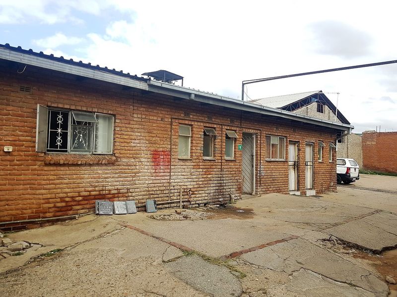 Rear exterior view of a commercial brick building featuring metal security bars, corrugated roofing, and a weathered concrete yard area.