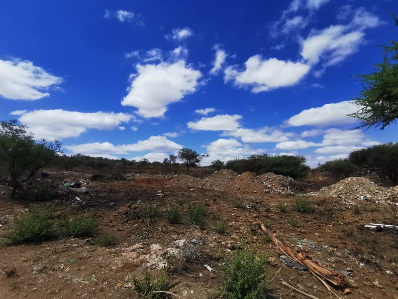 An undeveloped commercial land plot featuring natural terrain, native vegetation, and scattered debris under a clear blue sky.