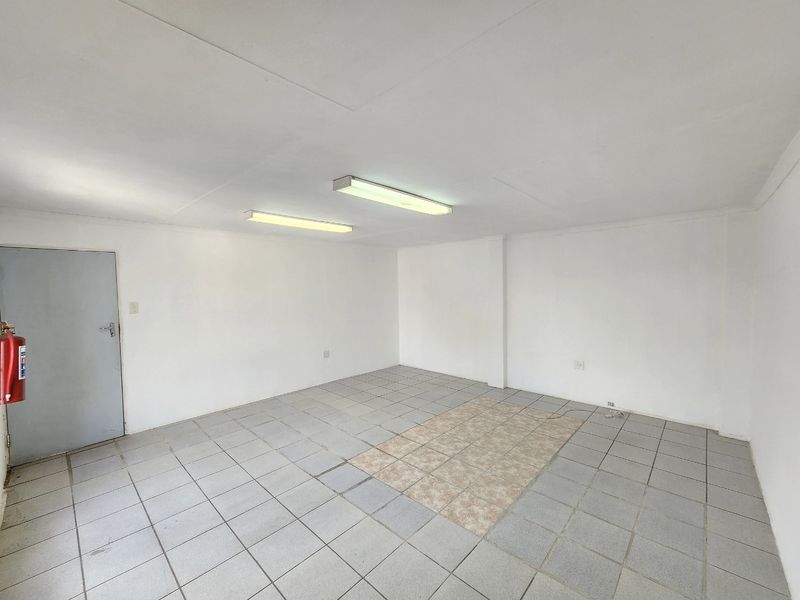 A vacant commercial office space featuring neutral white walls, tiled flooring with a central accent section, and overhead fluorescent lighting.
