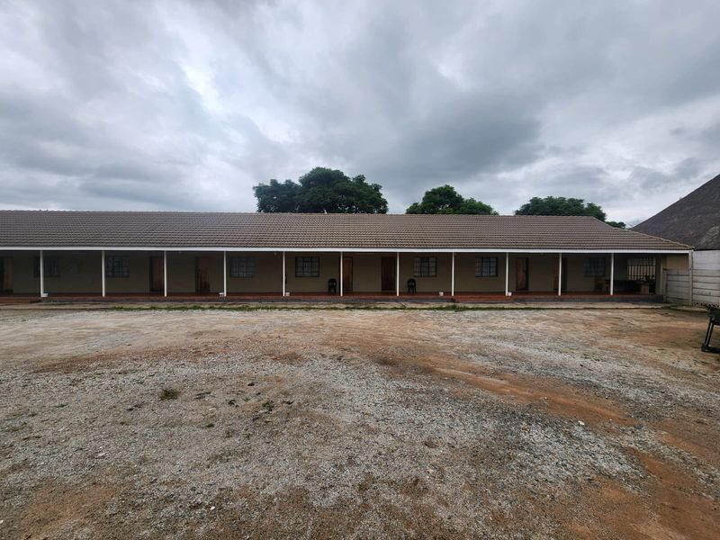 A long, single-story building featuring a covered veranda with multiple access points, tiled roofing, and a large, unpaved gravel yard area.