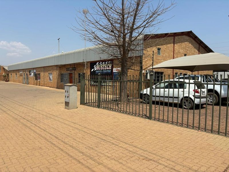 Front exterior view of an industrial warehouse facility featuring face brick construction, metal roofing, secure perimeter fencing, and a paved driveway with carport parking.