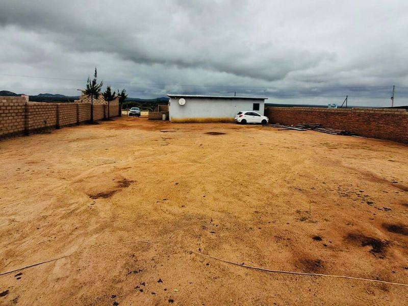A large, open, unpaved commercial plot enclosed by perimeter brick walls, featuring a small outbuilding structure.