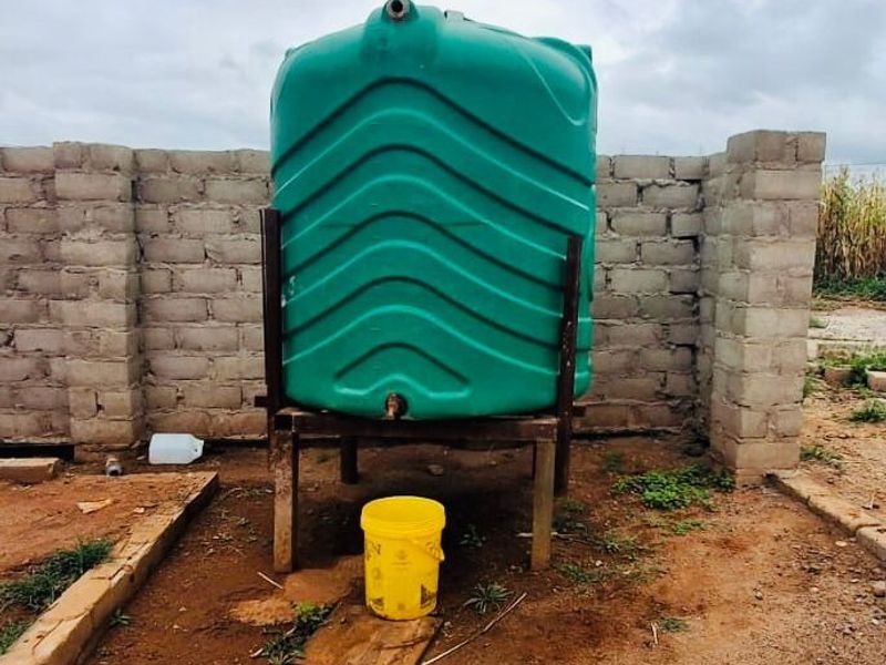 An outdoor utility area featuring a green plastic water storage tank mounted on a metal stand, enclosed by a cinder block wall.