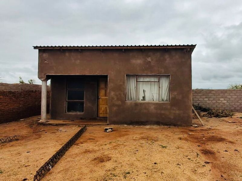 Front view of a single-story residential structure with unfinished plaster walls, metal roofing, and a surrounding brick boundary wall on a dirt lot.