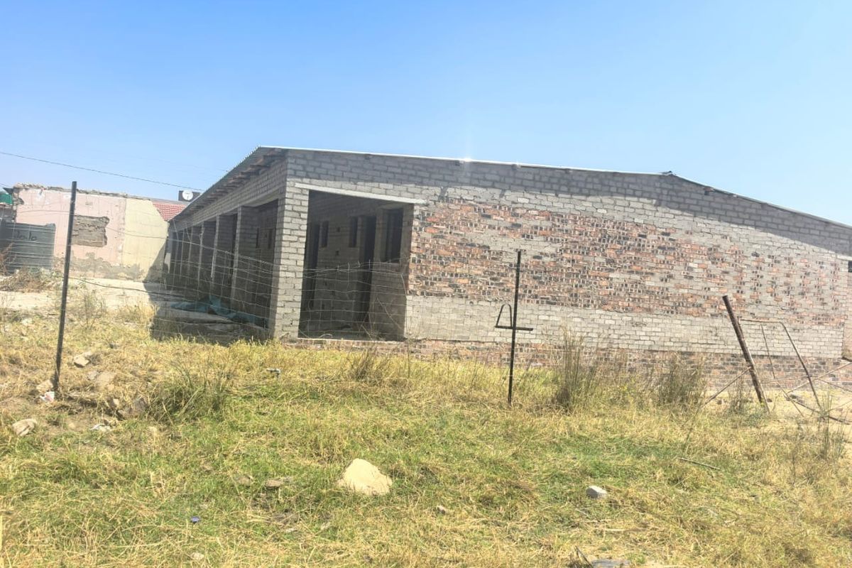 An unfinished exterior view of a residential building structure featuring exposed brick and concrete block walls with a corrugated metal roof.